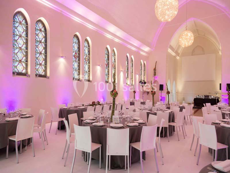 Salle de réception lumineuse avec tables rondes dressées, chaises blanches et vitraux colorés sur les murs.