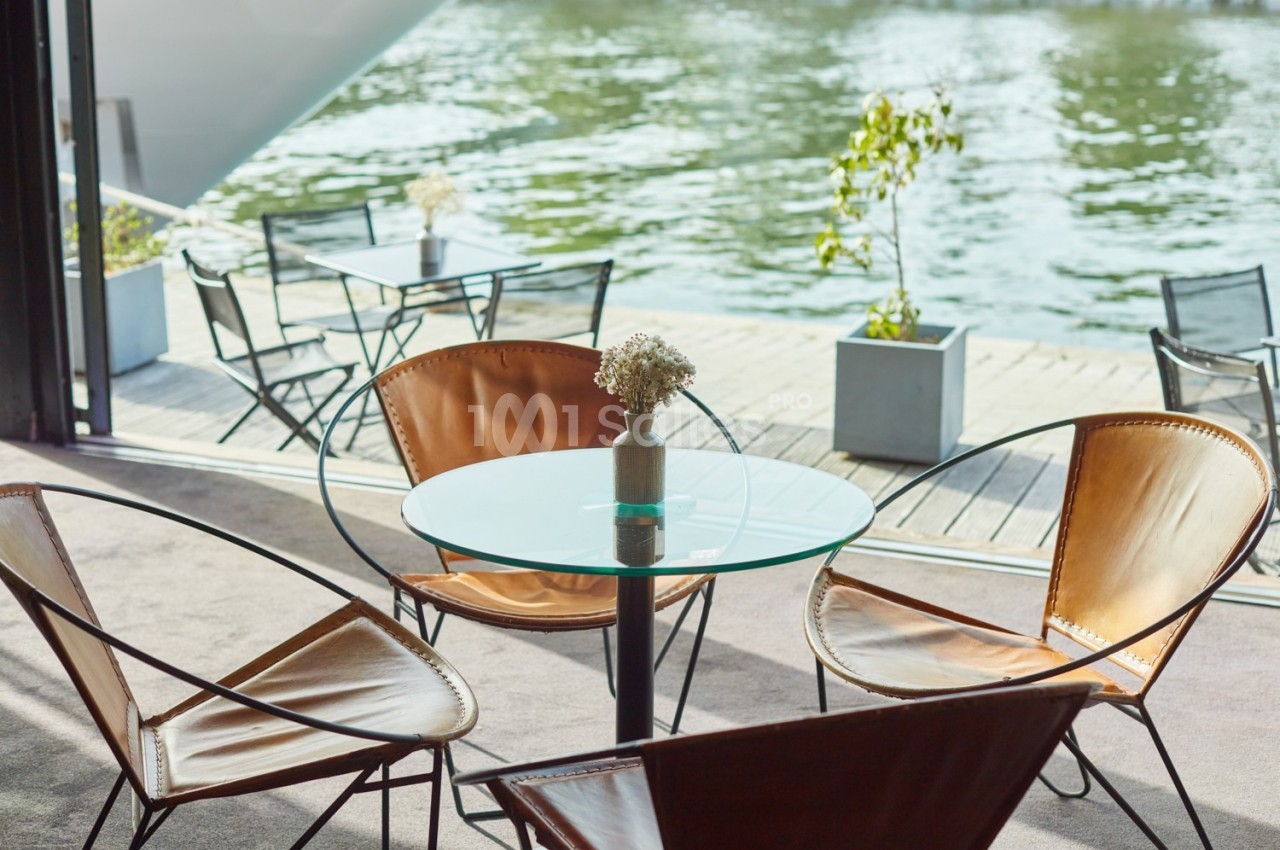 Table ronde en verre entourée de fauteuils en cuir, placée près d'une terrasse donnant sur un plan d'eau.
