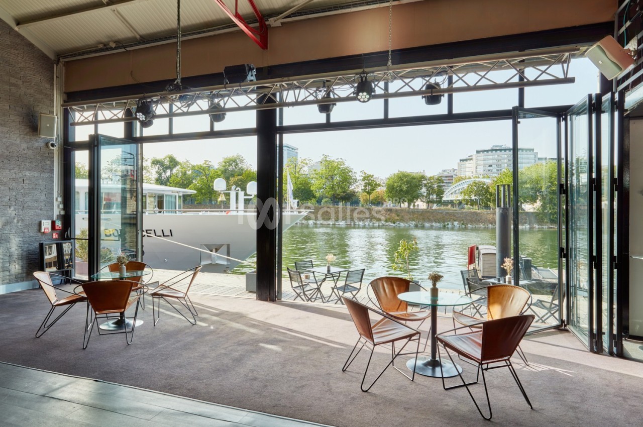 Salle lumineuse avec baies vitrées ouvertes sur une terrasse au bord de l'eau, aménagée avec tables et chaises modernes.