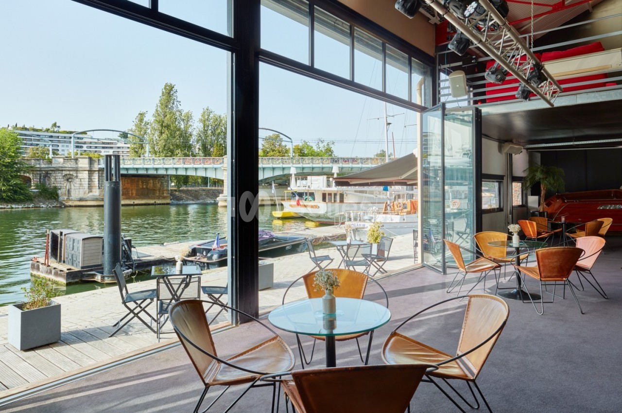 Salle avec baies vitrées donnant sur un quai, tables en verre et chaises design, vue sur un pont et des bateaux.