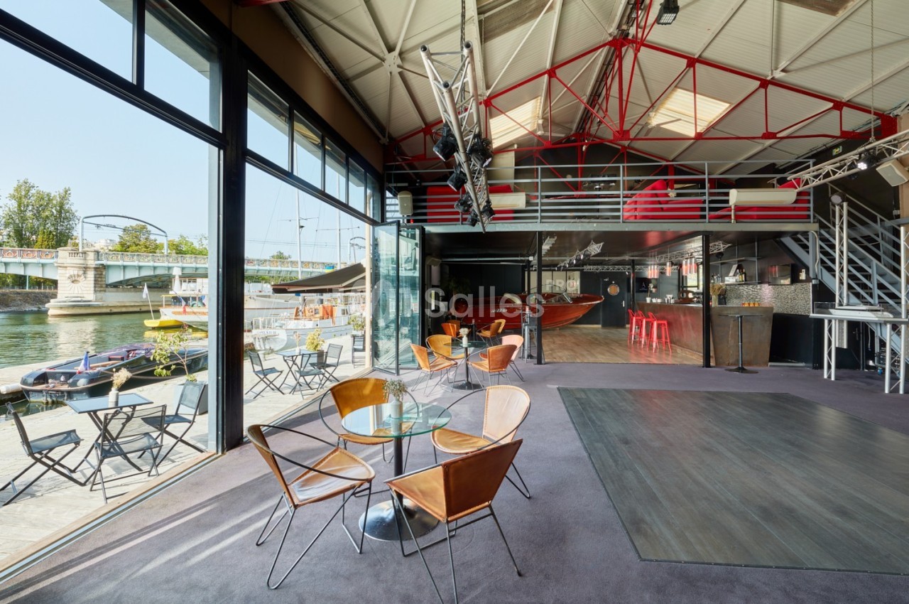 Salle lumineuse avec verrière, mobilier moderne, vue sur un quai et un pont, ambiance industrielle et conviviale.