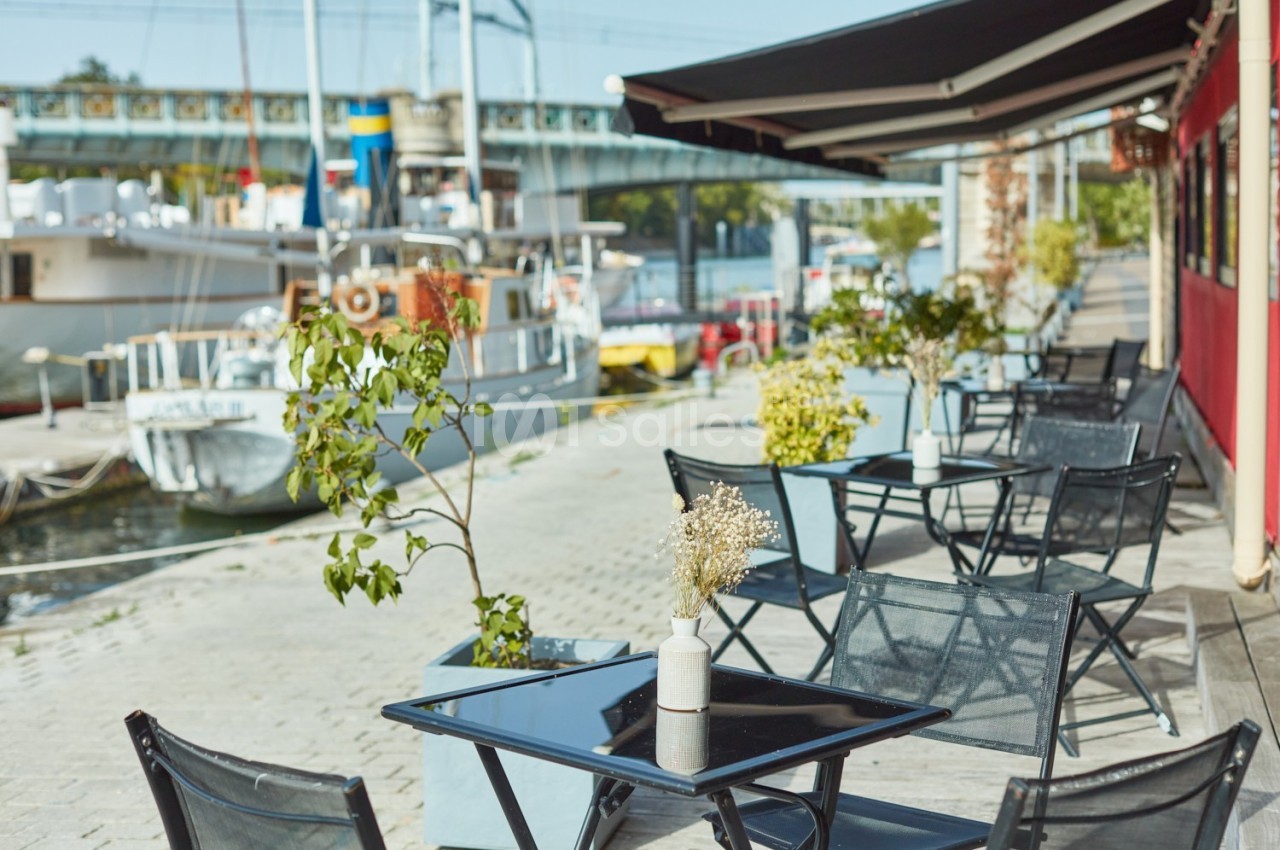 Terrasse avec tables et chaises noires, bordée de plantes, située le long d'un quai avec des bateaux amarrés.