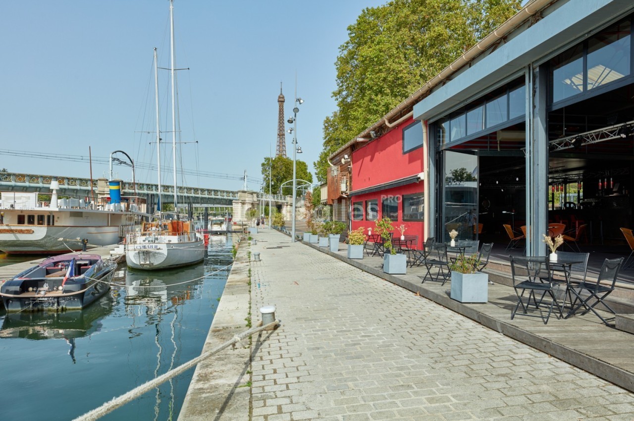 Terrasse avec tables et chaises le long d'un quai, près de bateaux amarrés et d'un bâtiment rouge, sous un ciel dégagé.