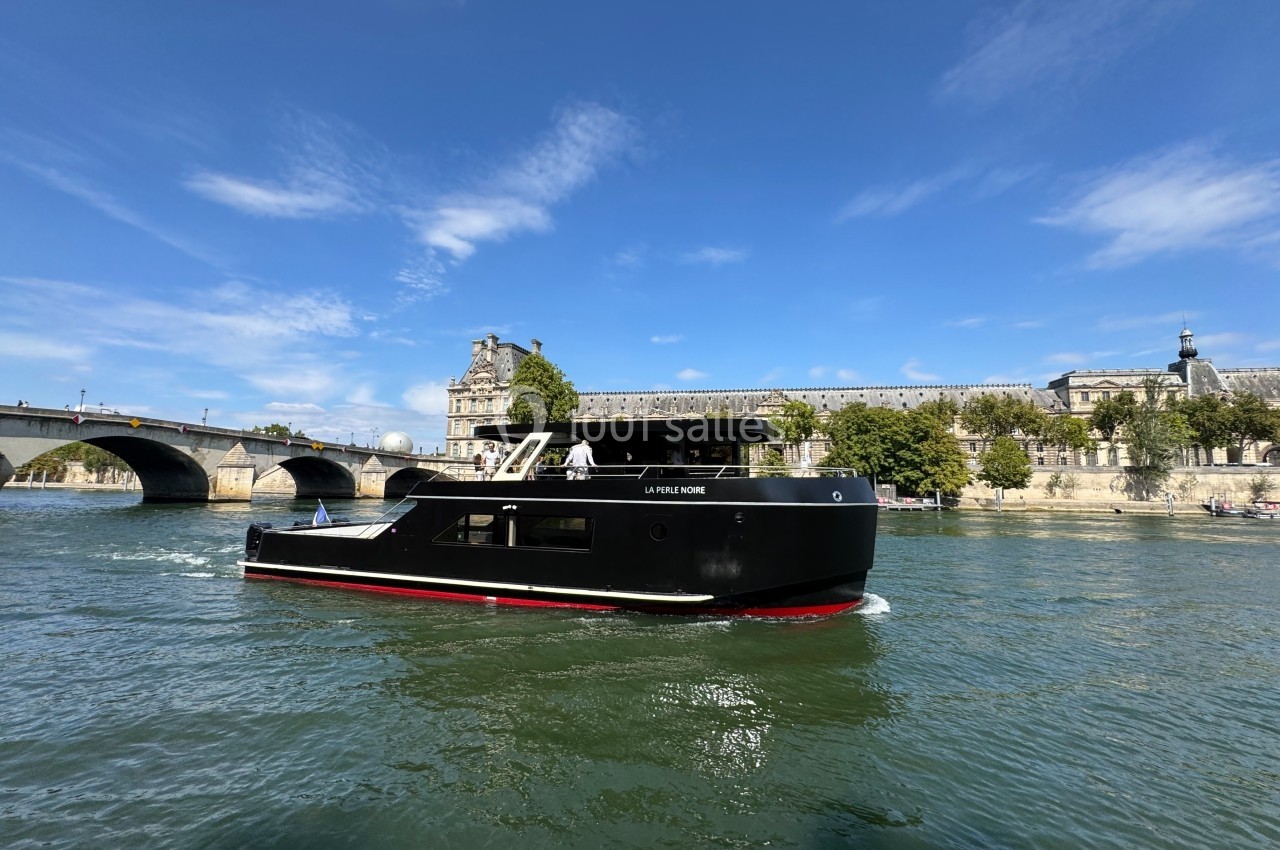 Bateau noir naviguant sur une rivière, avec un pont et des bâtiments historiques en arrière-plan sous un ciel bleu.