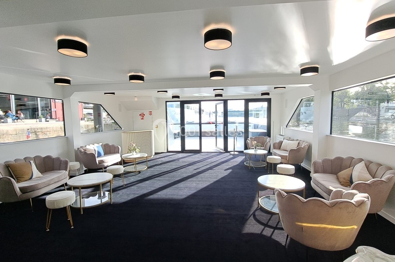 Salon lumineux avec canapés et tables basses, grandes baies vitrées offrant une vue sur l'eau et l'extérieur.