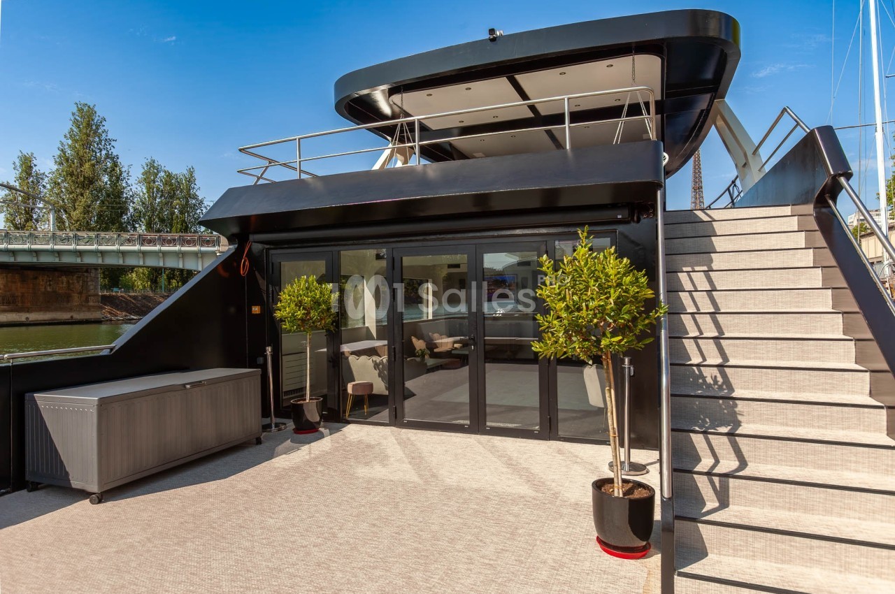Terrasse d'un bateau moderne avec escalier, plantes en pot et grandes baies vitrées donnant sur un intérieur lumineux.