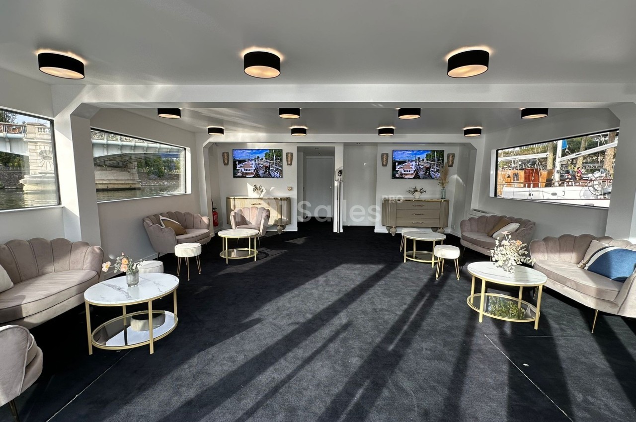 Salle lumineuse avec canapés, tables basses et décorations florales, offrant une vue sur l'extérieur par de grandes fenêtres.