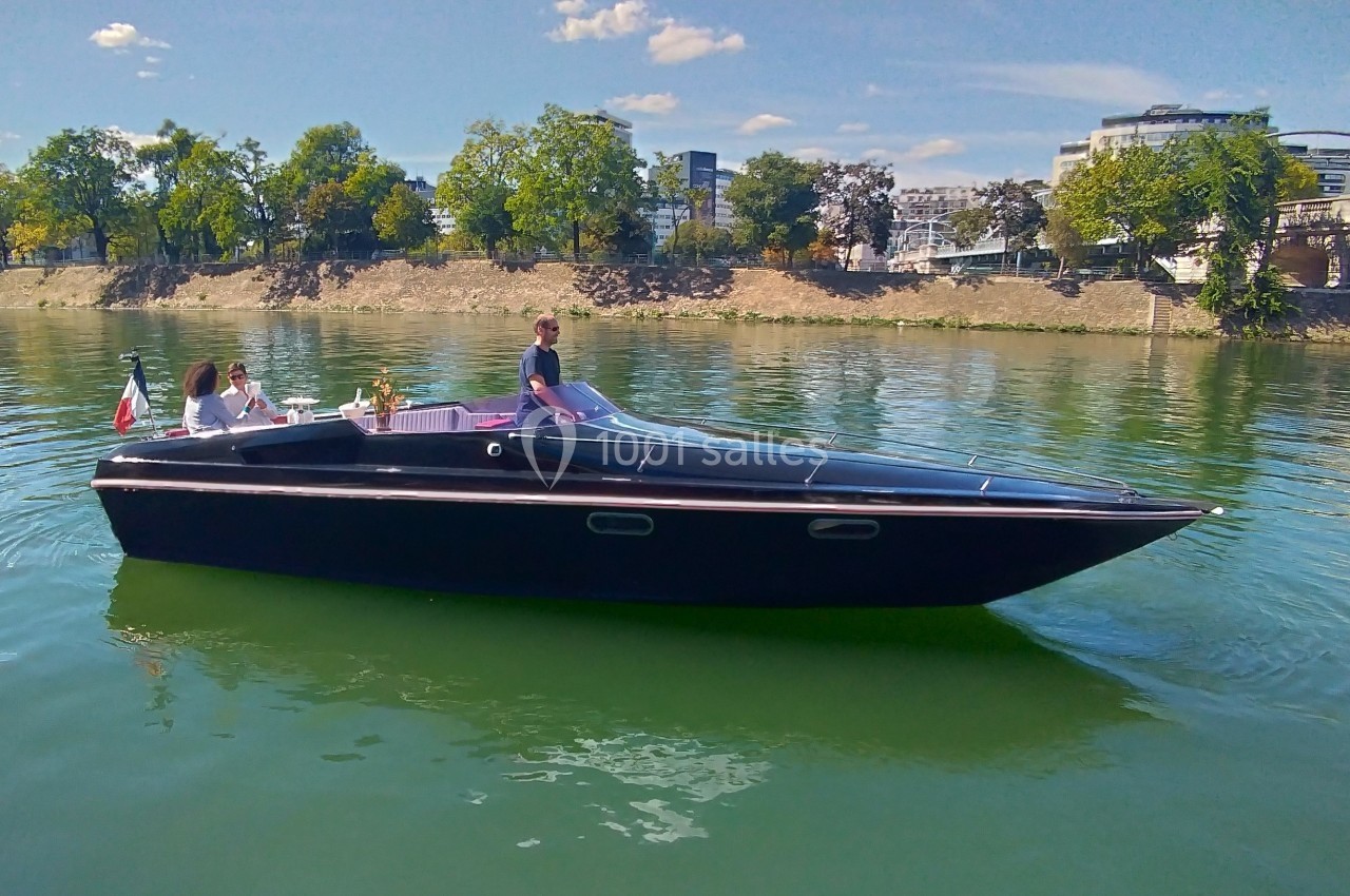 Bateau noir élégant naviguant sur une rivière bordée d'arbres et de bâtiments sous un ciel dégagé.