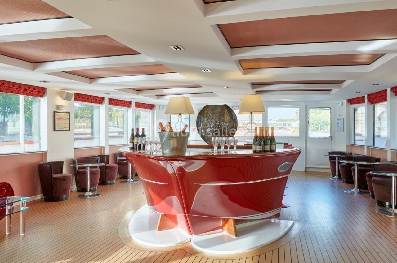 Bar moderne avec comptoir rouge brillant, bouteilles de champagne et verres, entouré de sièges dans une salle lumineuse.