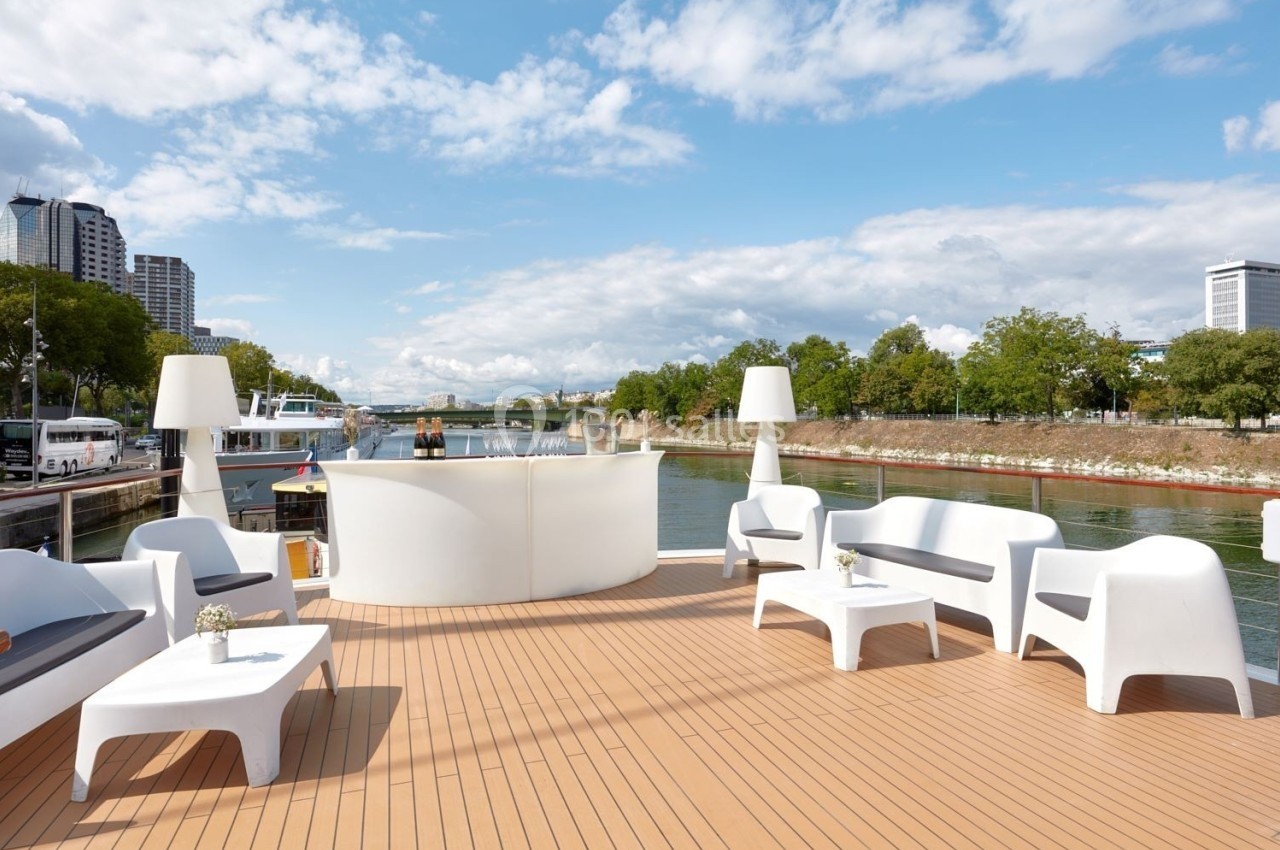 Terrasse d'un bateau avec mobilier moderne, vue sur un fleuve bordé d'arbres et bâtiments en arrière-plan.