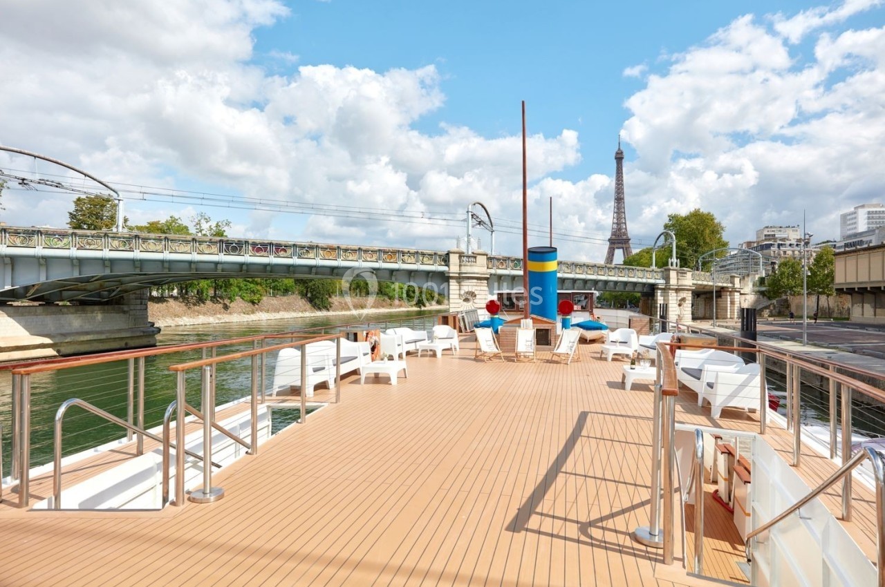 Pont supérieur d'un bateau avec des chaises longues, vue sur la Seine, un pont ferroviaire et la tour Eiffel en arrière-plan.