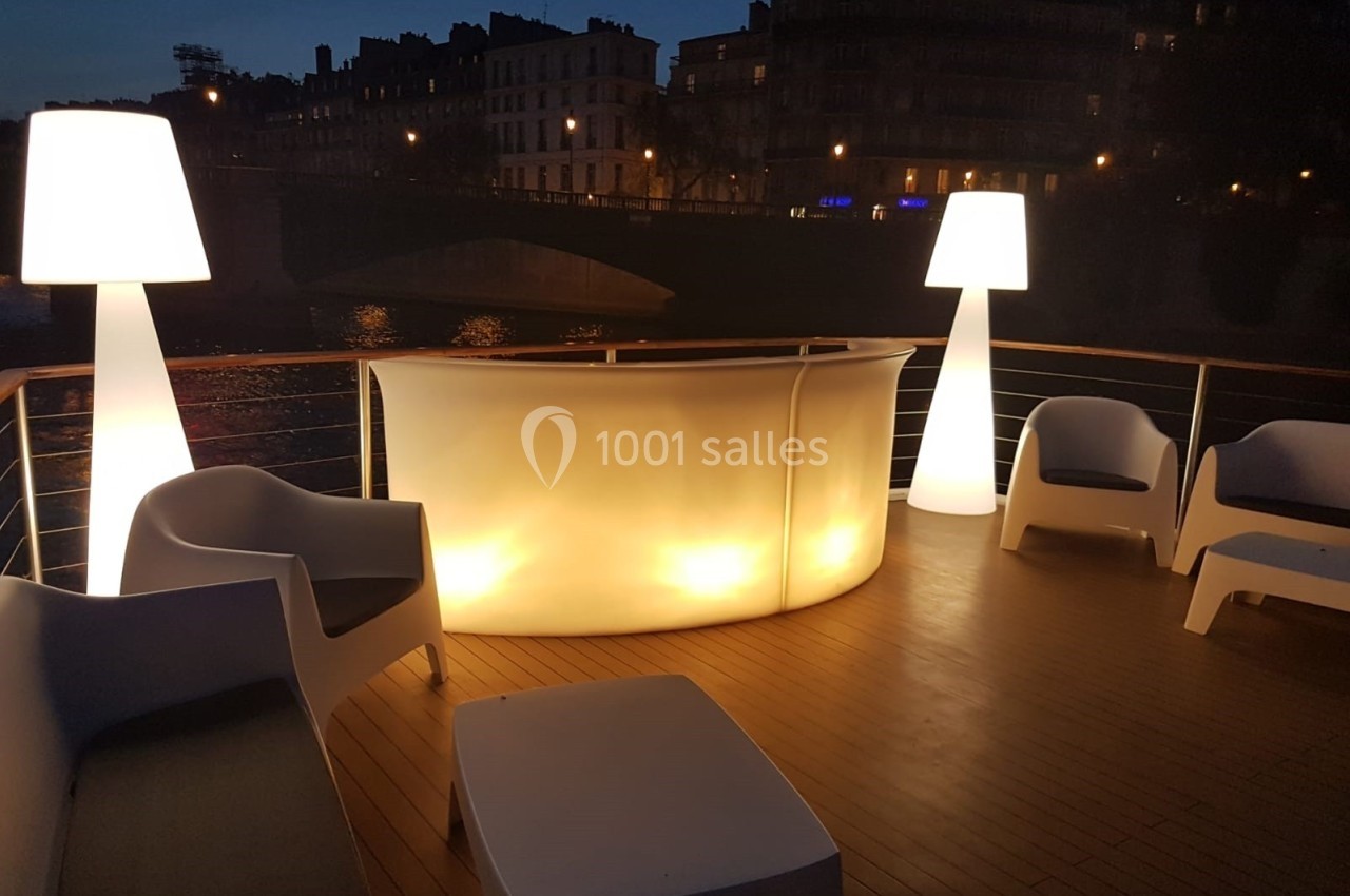 Terrasse en bois éclairée de nuit avec un bar lumineux, des fauteuils blancs et des lampes design près d'une rivière.