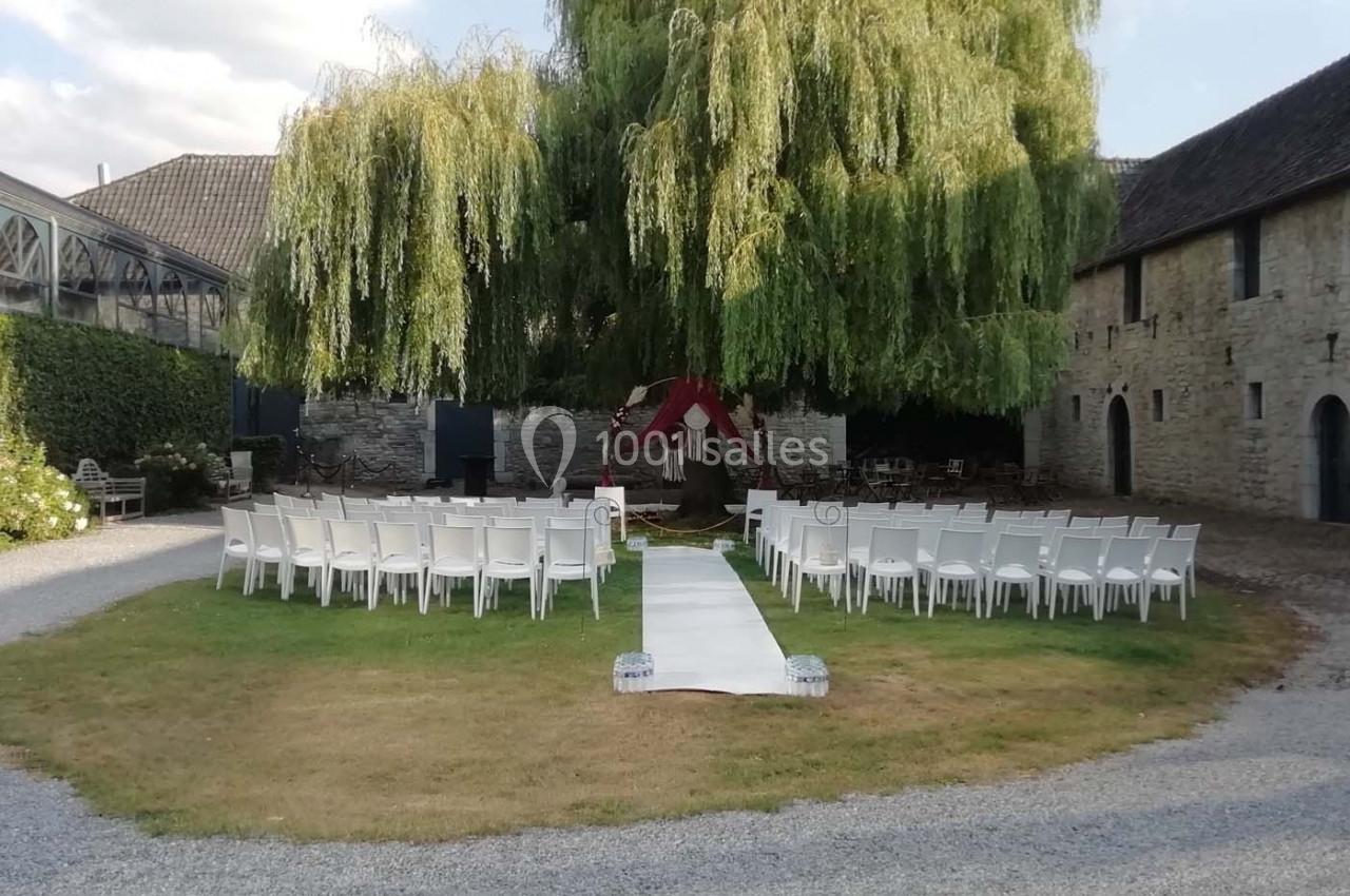 Chaises blanches disposées en arc autour d'une allée centrale devant un grand saule dans une cour en plein air.