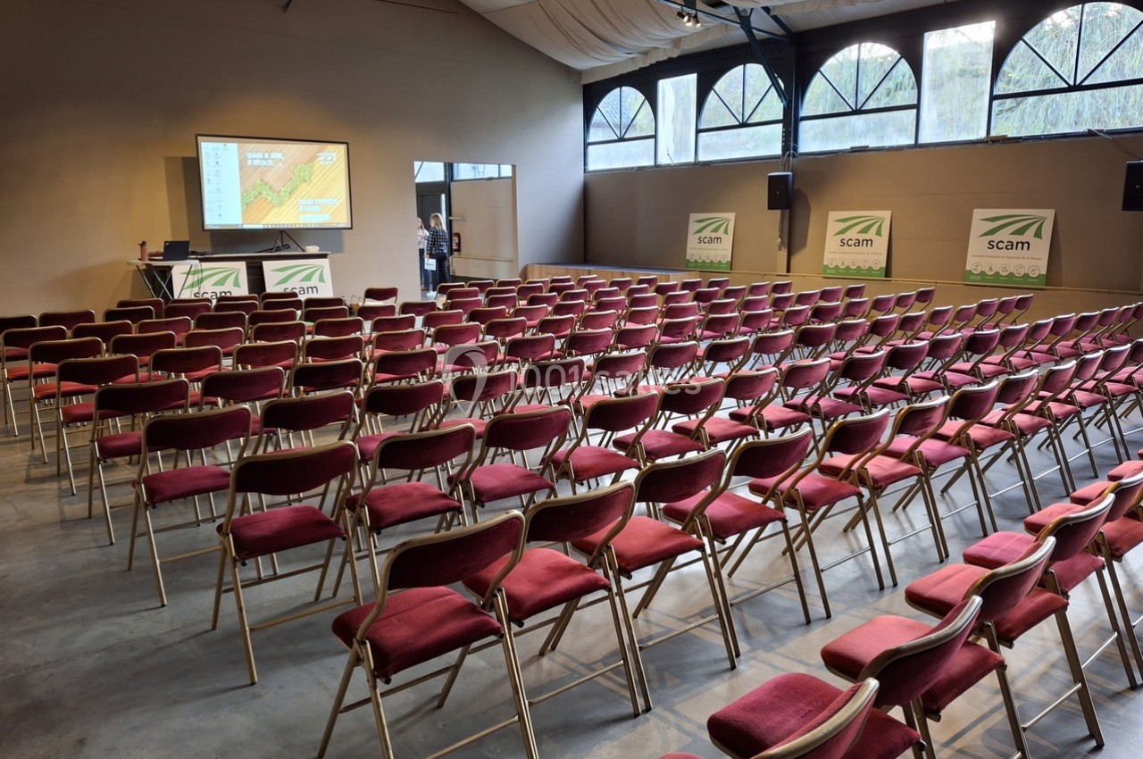 Salle de conférence avec des rangées de chaises rouges alignées, un écran de présentation allumé et des affiches murales.