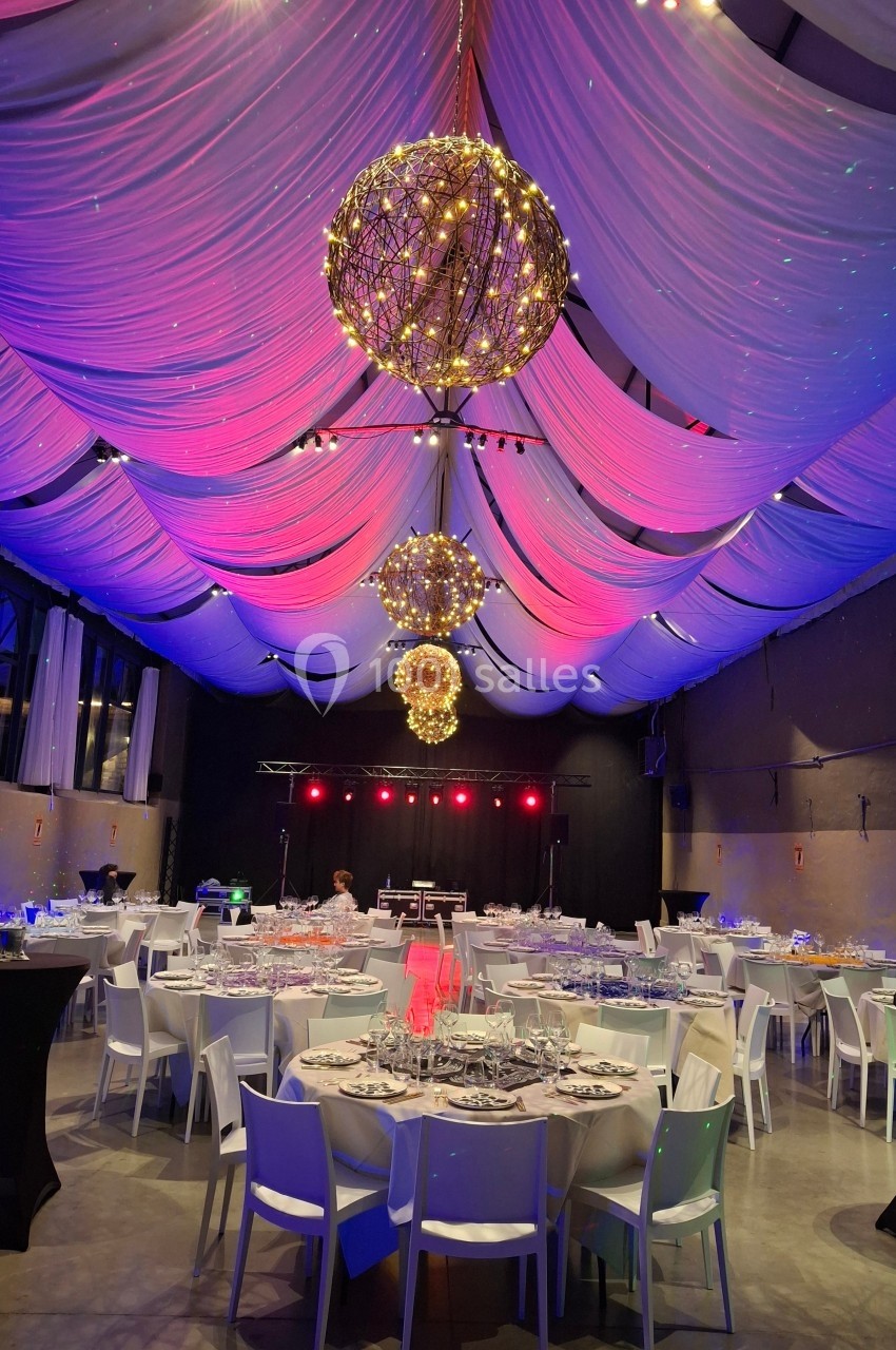 Salle de réception décorée avec des tables dressées, des guirlandes lumineuses et un éclairage coloré sous un plafond drapé.