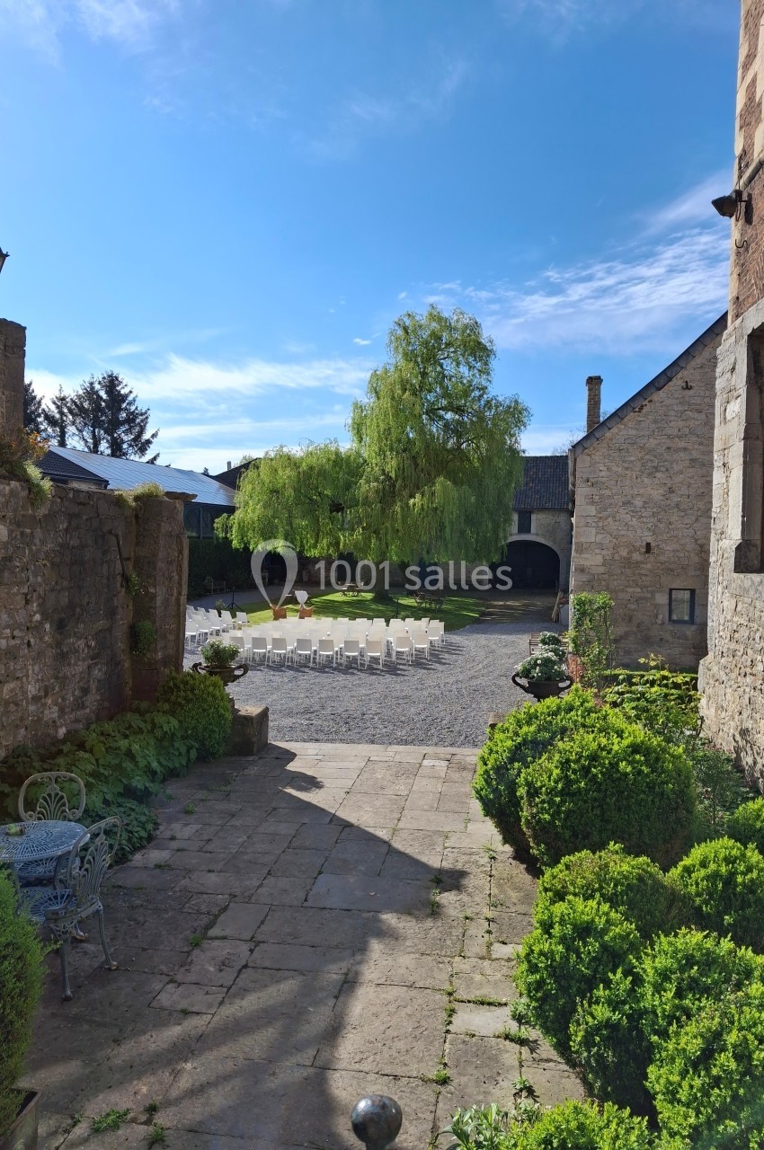 Cour pavée avec chaises blanches disposées en cercle, entourée de bâtiments en pierre et d'arbustes taillés.
