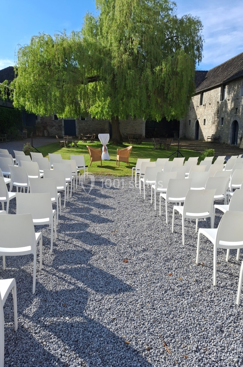Chaises blanches disposées en rangées face à un arbre et une table décorée dans une cour extérieure ensoleillée.