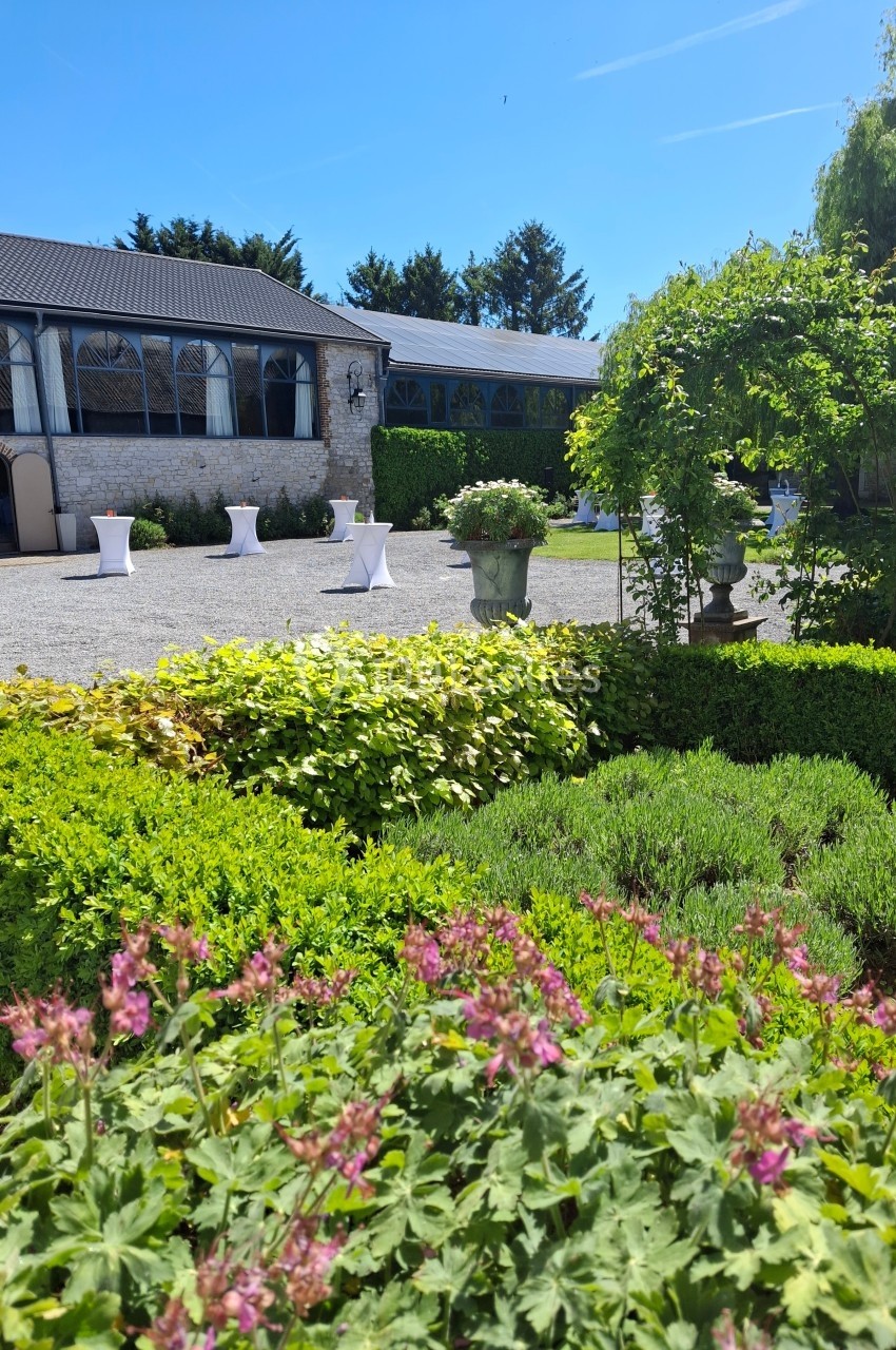 Jardin fleuri avec arbustes et parterres devant une terrasse pavée et un bâtiment en pierre sous un ciel bleu.
