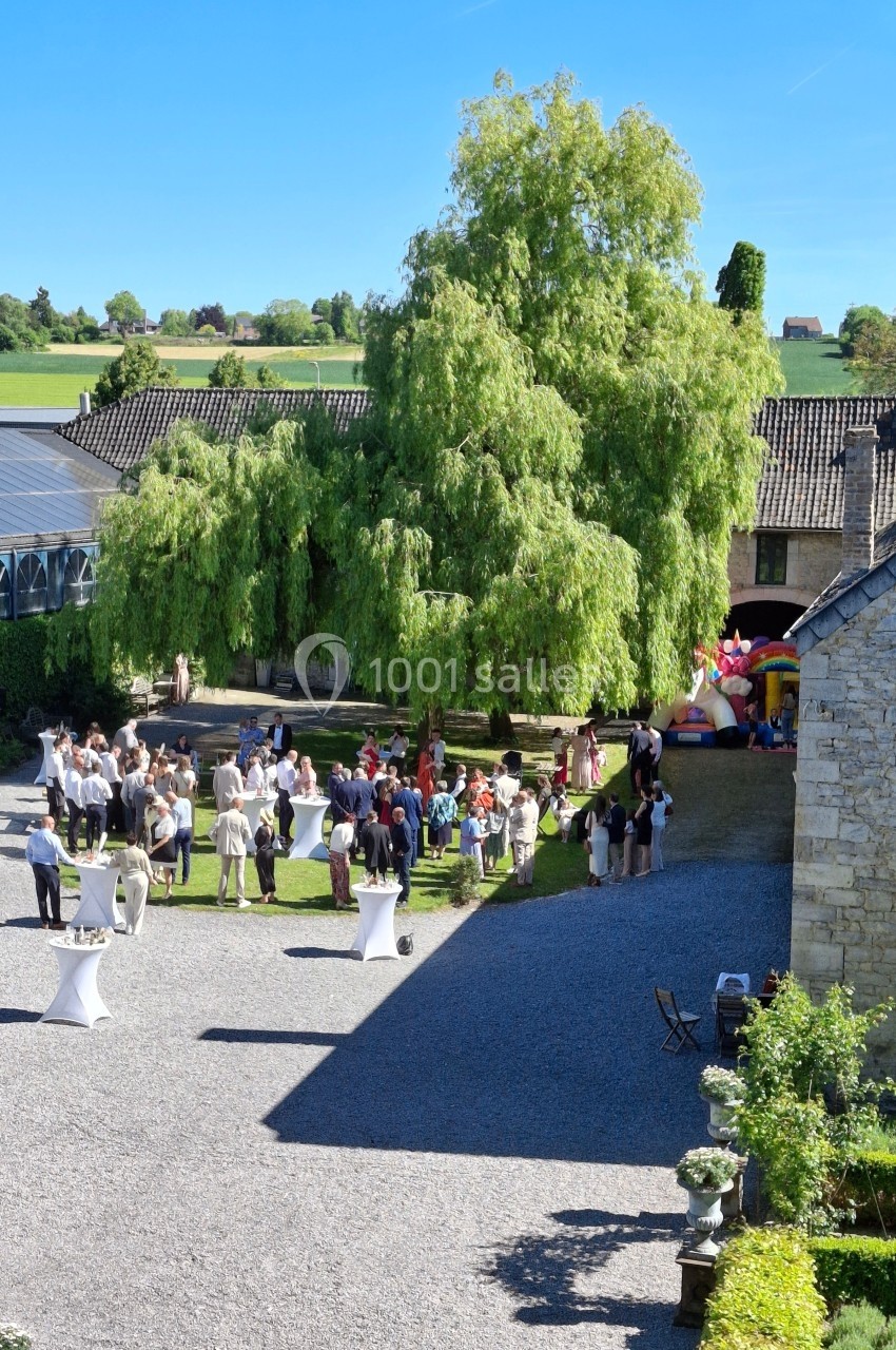 Groupe de personnes rassemblées dans une cour extérieure ensoleillée, entourée de bâtiments en pierre et de verdure.