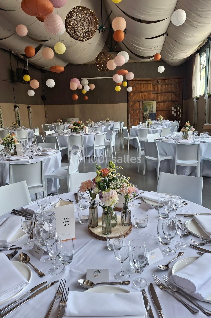 Salle de réception décorée avec des tables rondes dressées, des fleurs et des lanternes suspendues au plafond.