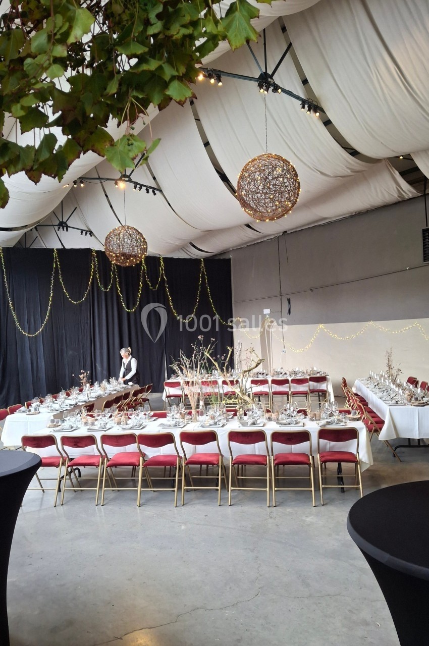 Salle de réception décorée avec des tables dressées, chaises rouges, guirlandes lumineuses et plafonds drapés blancs.