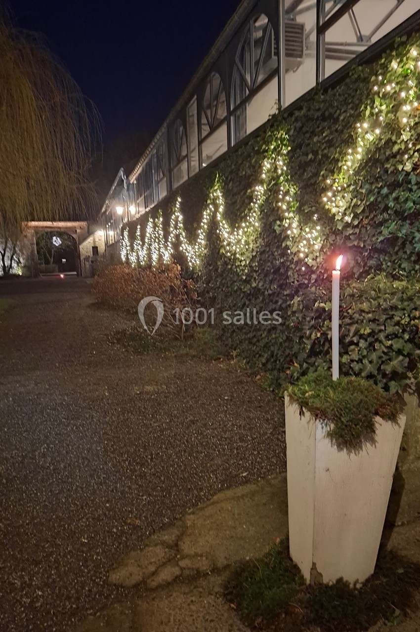 Mur végétal décoré de guirlandes lumineuses, allée pavée et bougie dans un pot blanc au premier plan, ambiance nocturne.
