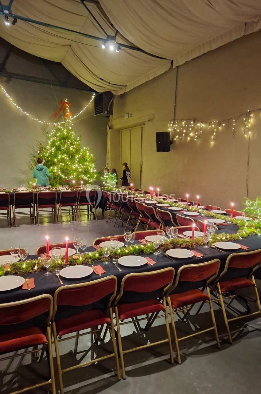 Salle décorée pour un repas festif avec longues tables dressées, guirlandes lumineuses et grand sapin illuminé.