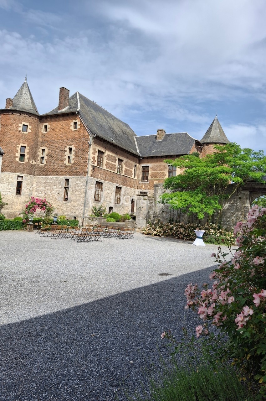 Cour d’un château en pierre et briques rouges, avec tours, fleurs, mobilier extérieur et ciel partiellement nuageux.