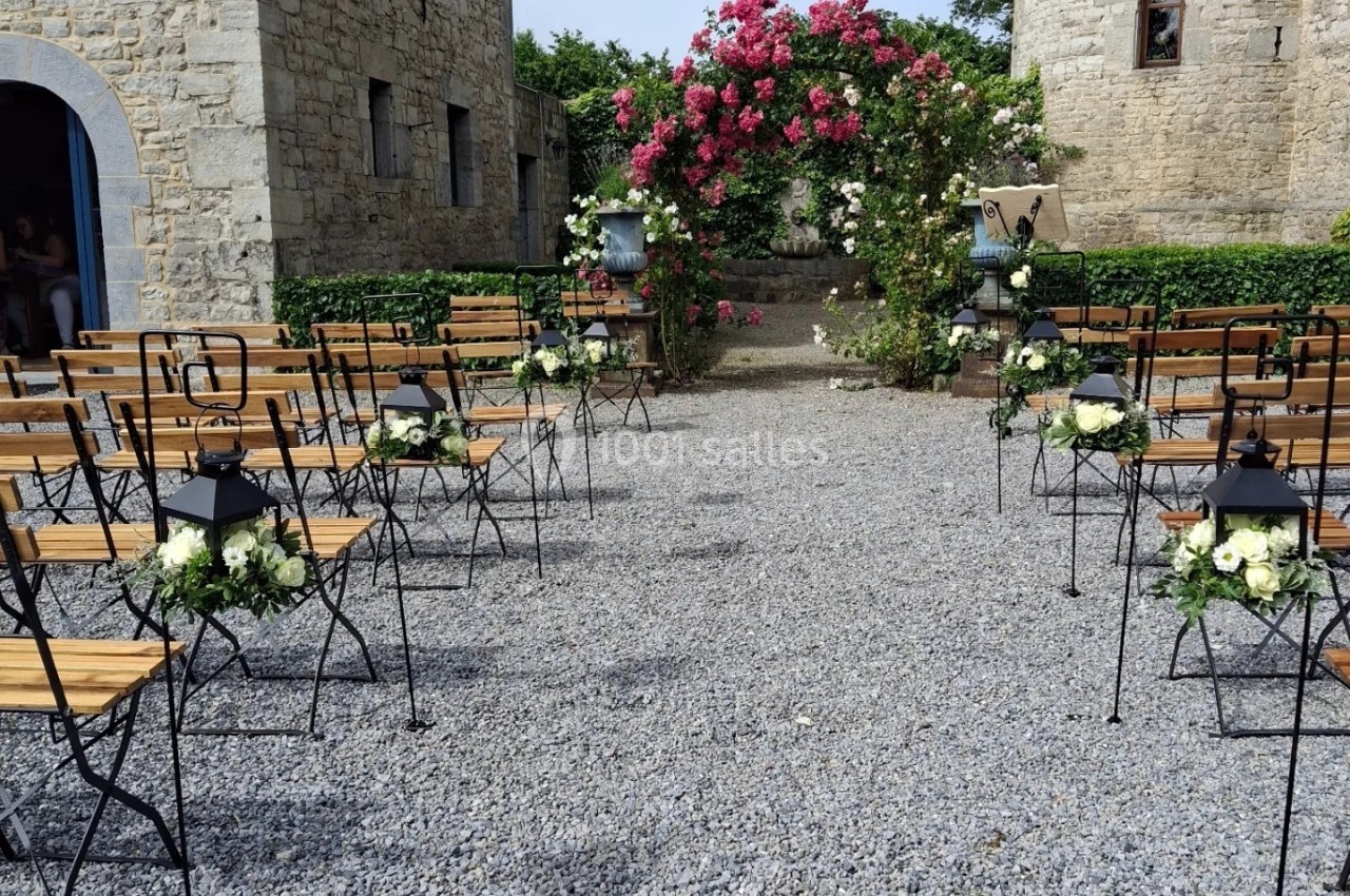 Allée extérieure décorée pour une cérémonie, avec bancs en bois, lanternes et fleurs blanches, devant une arche fleurie.