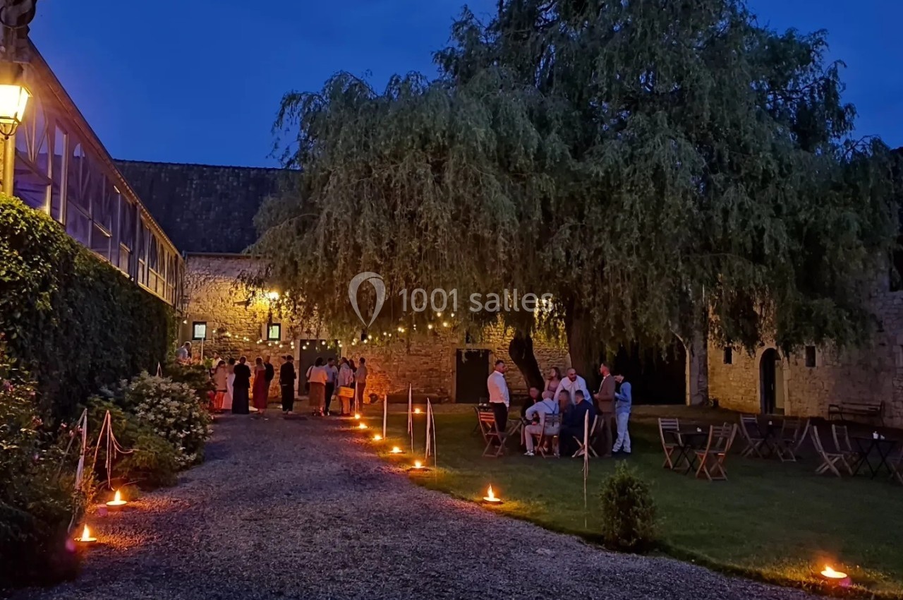 Cour extérieure éclairée par des lanternes, avec des invités rassemblés près d'un bâtiment en pierre au crépuscule.