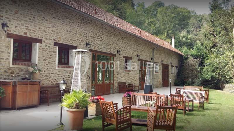 Façade en pierre d'un bâtiment avec terrasse aménagée, mobilier en bois et végétation environnante.