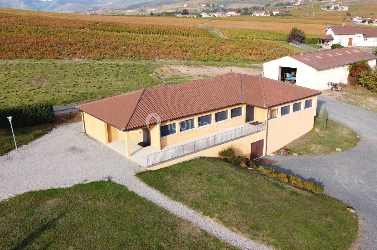 Bâtiment beige à toit rouge entouré de vignes et de champs, avec un chemin en gravier menant à l'entrée.