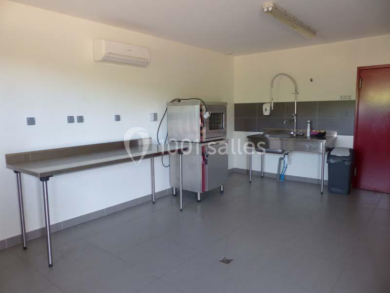 Cuisine professionnelle vide avec plan de travail en inox, évier, micro-ondes et sol carrelé gris.