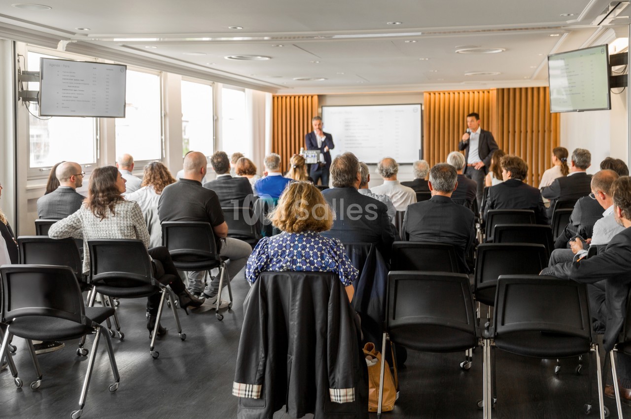 Un groupe de personnes assises dans une salle de conférence, écoutant deux intervenants devant un écran.