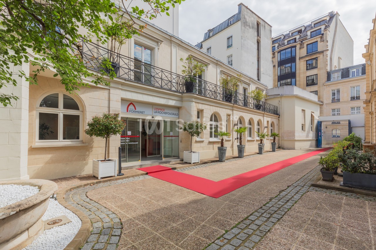 Entrée d'un bâtiment avec une façade claire, un tapis rouge au sol et des plantes en pots alignées.
