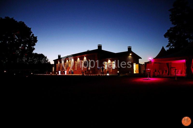 Bâtiment en pierre éclairé de nuit avec des lumières chaudes, entouré d'arbres et d'une tente illuminée.