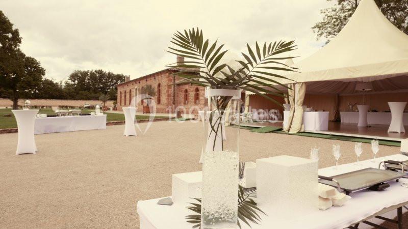 Décoration de table avec vase et palmes devant une tente blanche et un bâtiment en briques dans un espace extérieur.