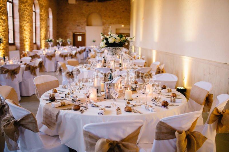 Salle de réception décorée pour un mariage, avec tables rondes ornées de nappes blanches, bougies et fleurs.