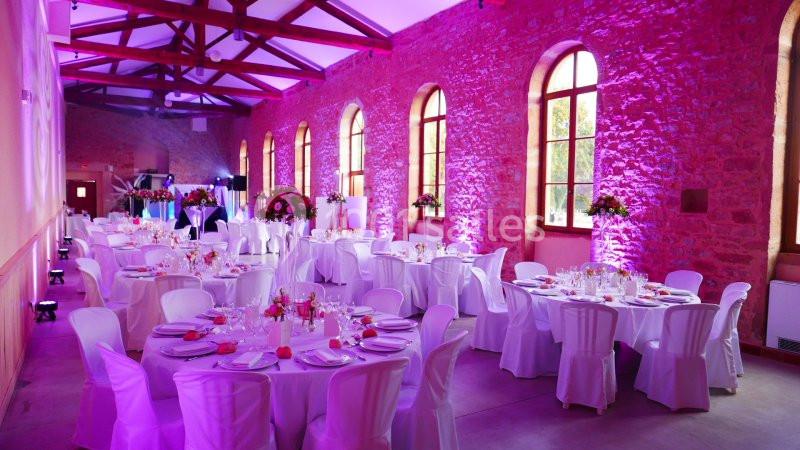 Salle de réception décorée avec des tables rondes, nappes blanches, chaises assorties et éclairage rose.