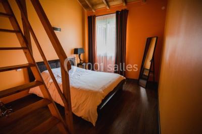 Chambre avec lit double, parquet clair, échelle en bois menant à une mezzanine, fenêtre avec rideaux et miroir debout.