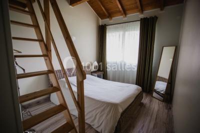Chambre avec lit double, parquet clair, échelle en bois menant à une mezzanine, fenêtre avec rideaux et miroir debout.