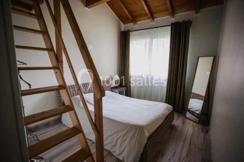Chambre avec lit double, parquet clair, échelle en bois menant à une mezzanine, fenêtre avec rideaux et miroir debout.