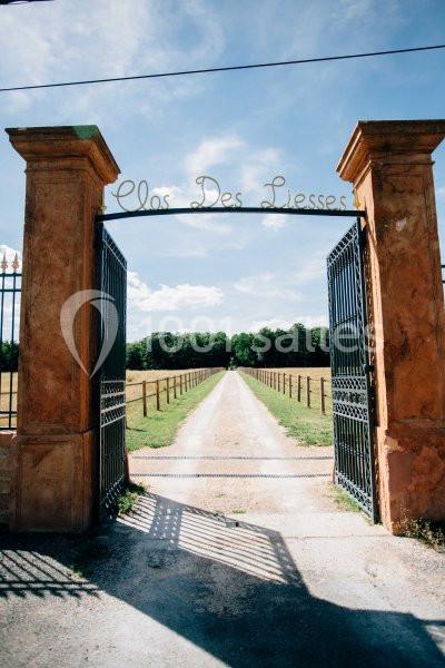 Entrée d'un domaine avec portail ouvert, chemin de gravier central et champs bordés de clôtures sous un ciel bleu.