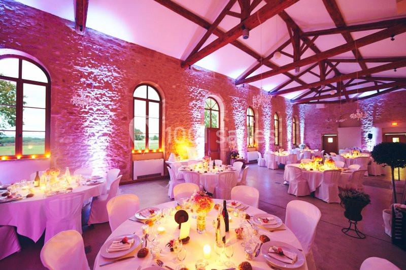 Salle de réception en pierre avec tables rondes décorées, éclairage coloré et grandes fenêtres en arc.