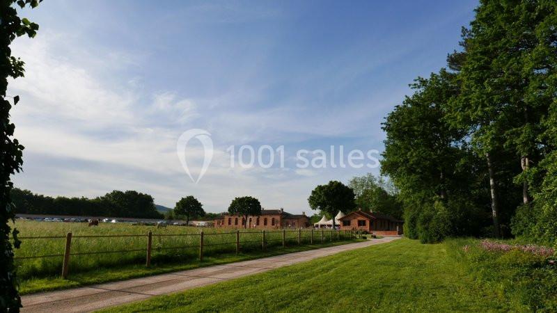 Chemin bordé de pelouse menant à des bâtiments en briques, entourés de prairies et d'arbres sous un ciel dégagé.