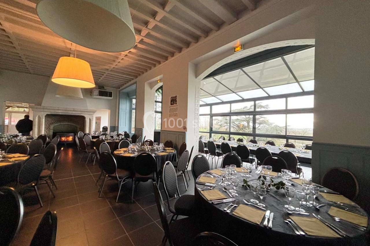 Salle de réception lumineuse avec tables rondes dressées, grandes fenêtres et cheminée en arrière-plan.