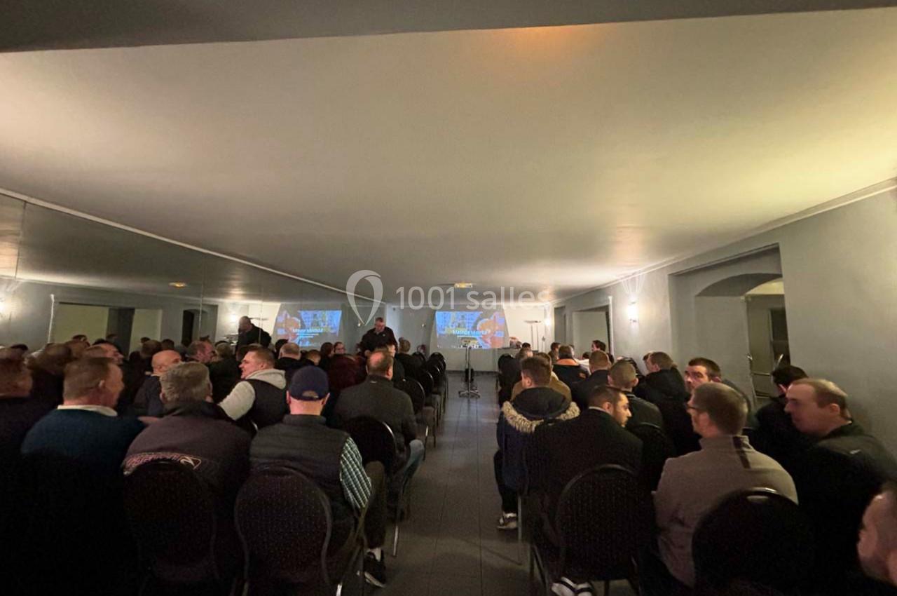 Salle remplie de personnes assises, regardant une présentation projetée sur deux écrans au fond de la pièce.