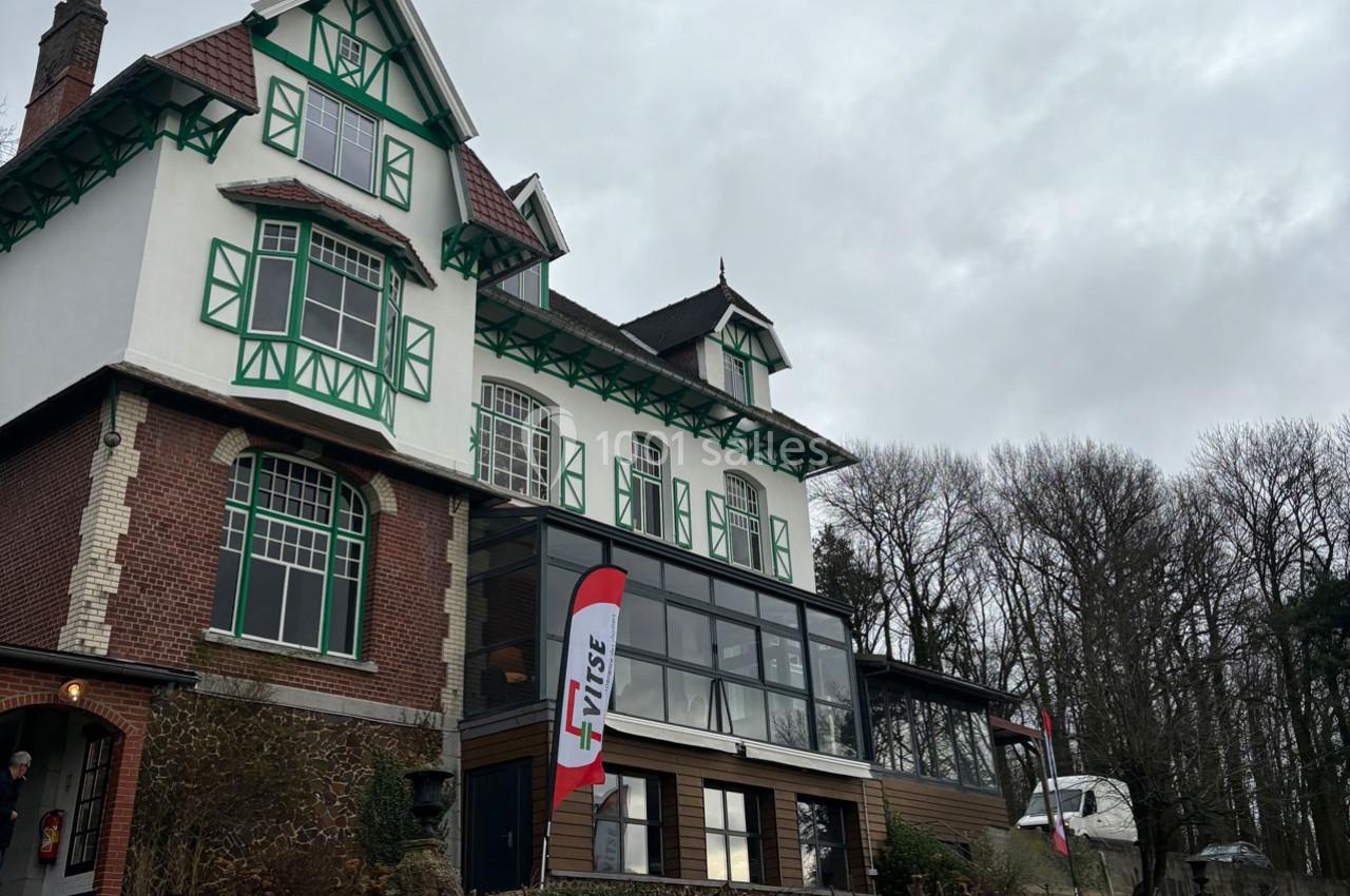 Façade d'une grande maison à colombages verts avec une extension vitrée, entourée d'arbres sous un ciel nuageux.