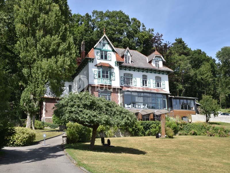 Grande maison à colombages entourée de verdure, avec un arbre taillé au premier plan et une forêt en arrière-plan.