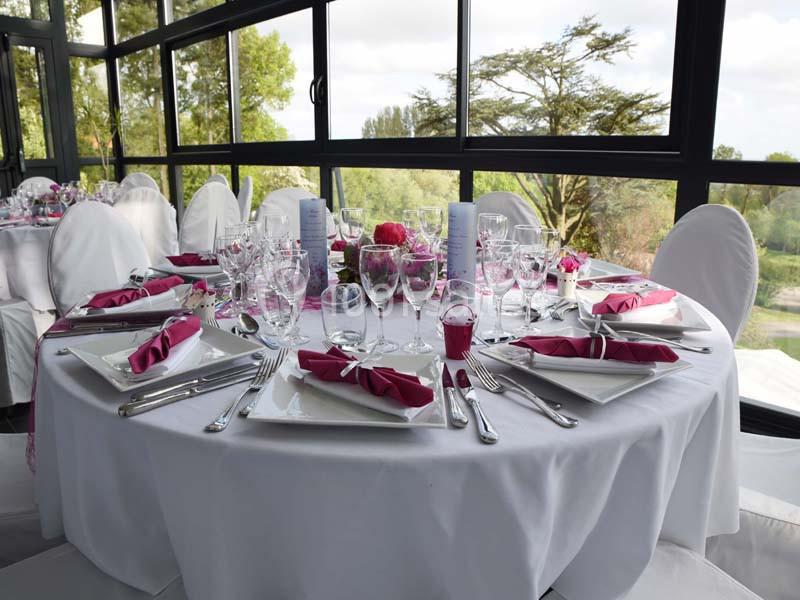Table ronde élégamment dressée avec nappes blanches, serviettes roses pliées et verrerie, devant de grandes baies vitrées.